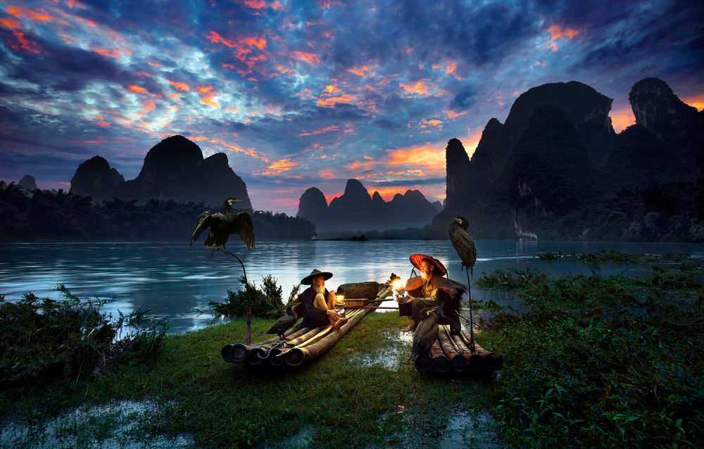 Обои для рабочего стола Китайские рыбаки сидят в лодках при свете фонаря на берегу горного озера, с ними рядом сидят птицы