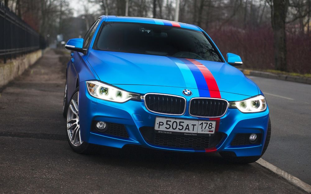 Обои для рабочего стола Голубой bmw 335i стоит на обочине возле забора на фоне деревьев