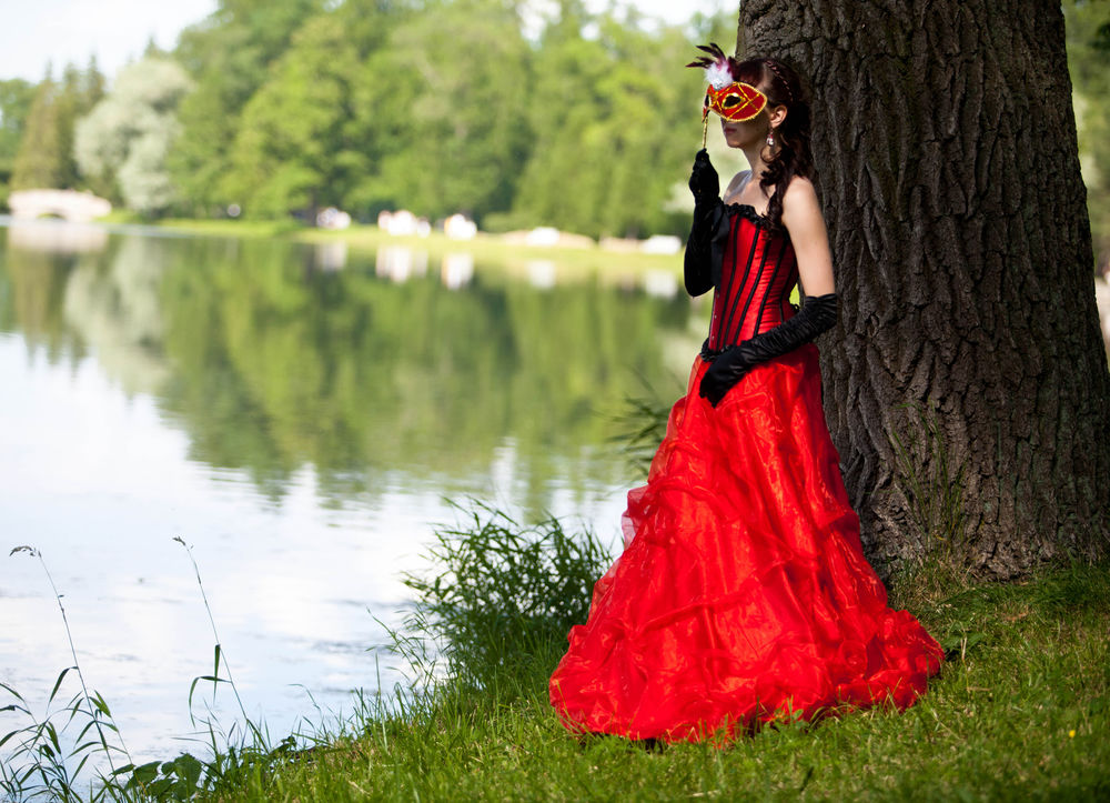 Обои для рабочего стола Девушка в красном платье с маской, стоит под деревом на берегу озера