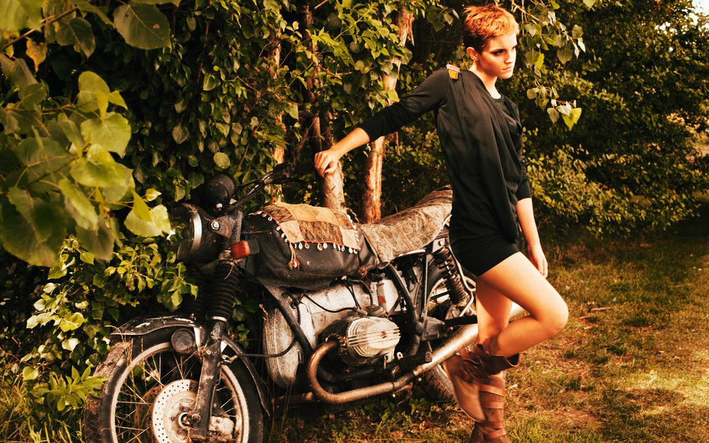 Обои для рабочего стола Актриса Emma Watson в черном платье позирует рядом с мотоциклом на фоне кустов и деревьев