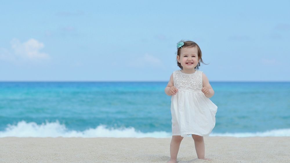 Обои для рабочего стола Смеющаяся девочка в белом платье стоит на песке возле моря