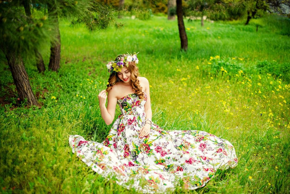 Обои для рабочего стола Девушка в венке из разноцветных цветов и в летнем платье с яркими цветами сидит на зеленой лужайке. Фотограф Анна Асланян