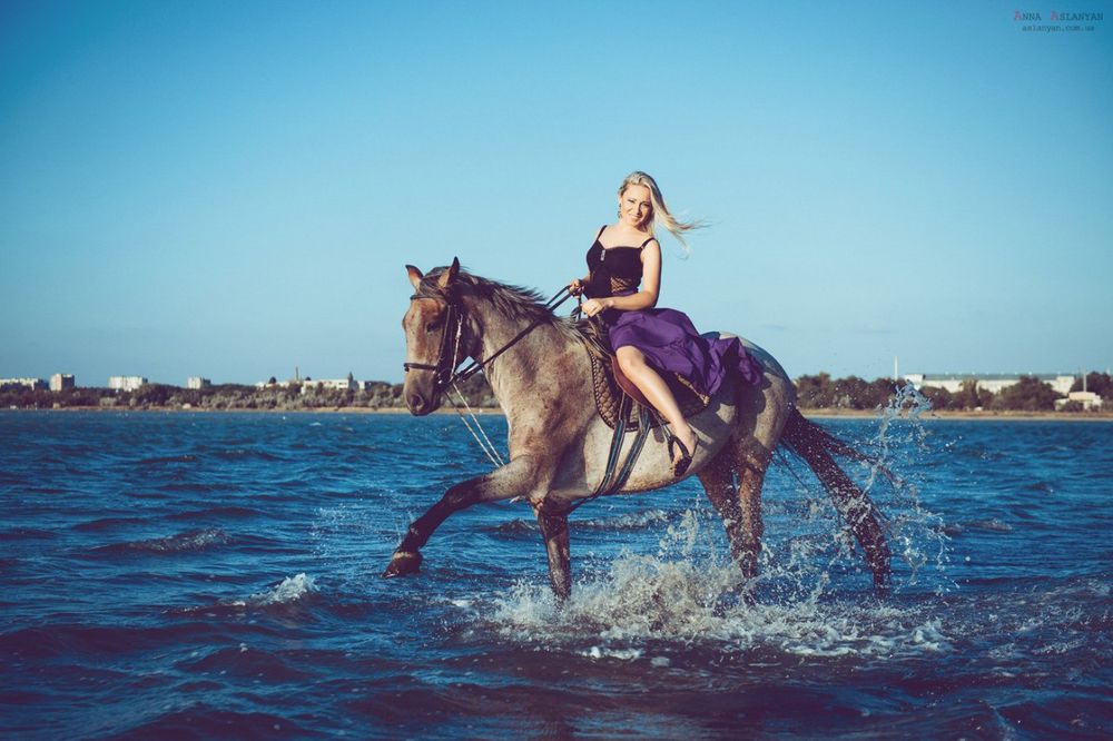 Обои для рабочего стола Девушка блондинка с длинными волосами едет верхом на лошади по прибрежной морской полосе. Девушка улыбается. Вдали видна береговая полоса со зданиями. Фотограф Анна Асланян