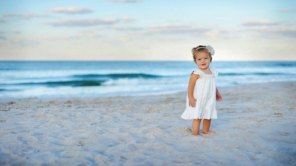 Обои для рабочего стола Малышка в белом платье с цветком на голове стоит в песке на берегу моря на фоне неба
