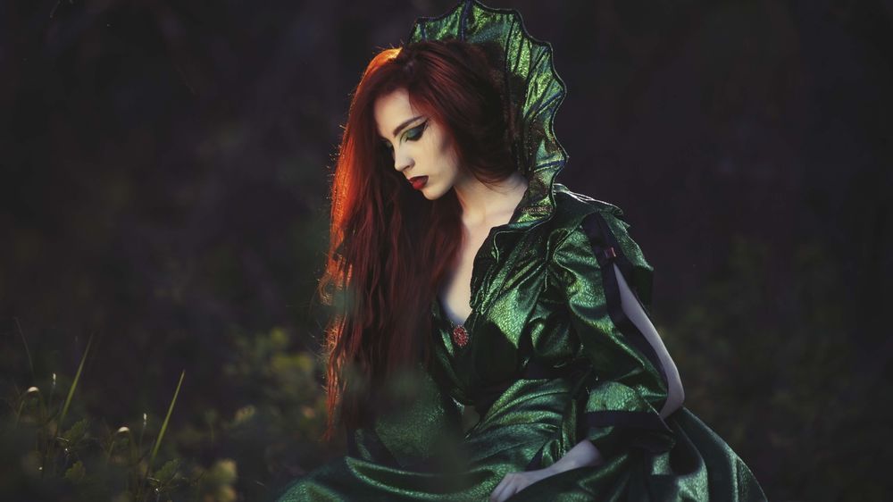 Обои для рабочего стола Рыжеволосая фея в зеленом платье сидит в лесу, опустив взгляд на землю, на темном фоне леса