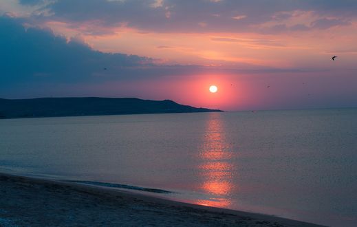 Обои на рабочий стол Закат солнца на фоне спокойного моря, на переднем плане и слева на фото видна береговая полоса