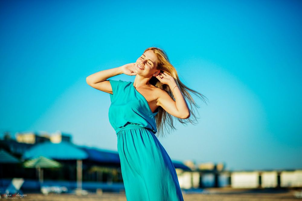 Обои для рабочего стола Девушка блондинка в голубом платье стоит на пляже, она закрыла глаза, завела руки за голову и улыбается солнцу. Фотограф Анна Асланян