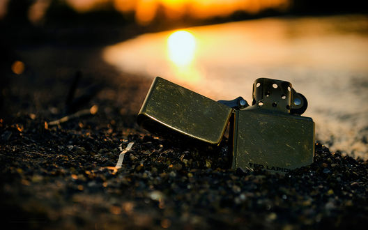 Обои на рабочий стол Зажигалка Zippo лежит на песчаном берегу, заходящее солнце отражается в воде