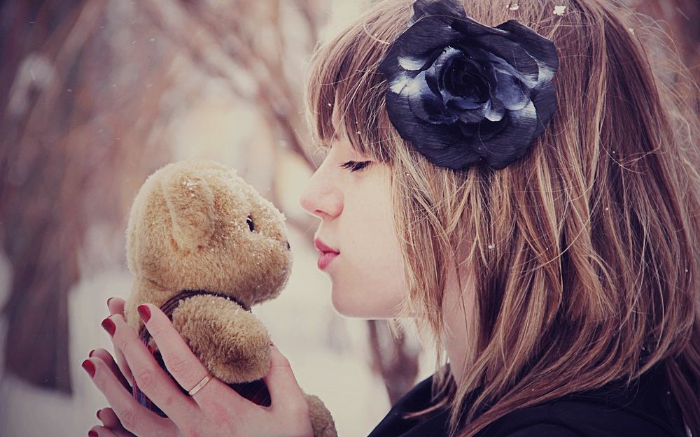 Обои для рабочего стола Девушка с заколкой в виде черной розы, держит в руках игрушечного медведя, хочет поцеловать медвежонка