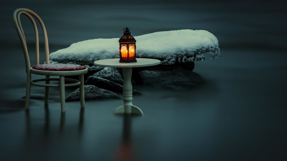 Обои для рабочего стола Камень, покрытый коркой снега и льда, стул и столик, на котором стоит горящий фонарь, стоят в водной глади