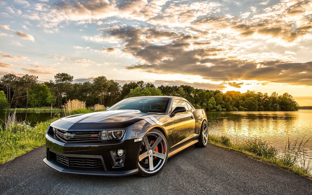 Обои для рабочего стола Chevrolet Camaro стоит на дороге, по бокам которой находится озеро. На заднем плане виднеется летний лес