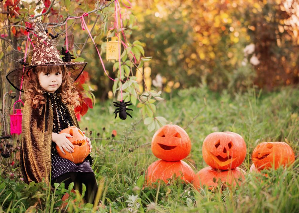 Обои для рабочего стола Маленькая девочка, нарядилась к Хэллоуину в костюм колдуньи, сидит около деревьев, держа тыкву в руках