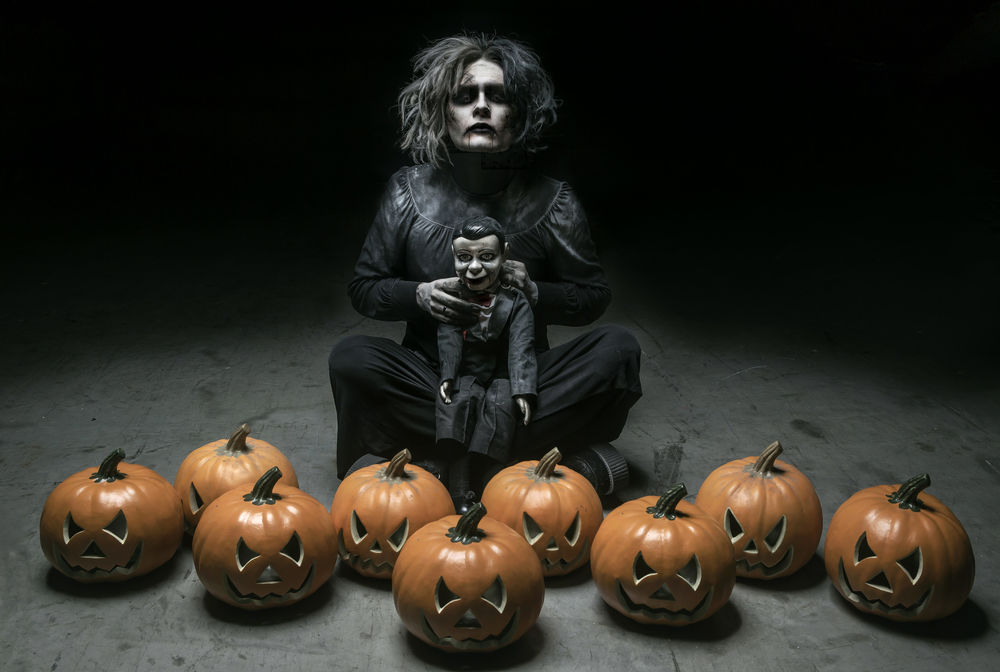 Обои для рабочего стола Мрачный мужчина с куклой в руках, сидит на полу в темном помещении при свете лампочки, среди тыкв на Хэллоуин