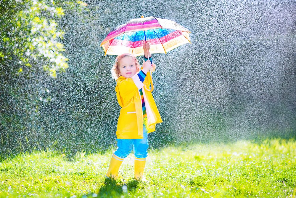 Обои для рабочего стола Маленькая, ярко одетая девочка стоит под зонтиком на лужайке во время дождя