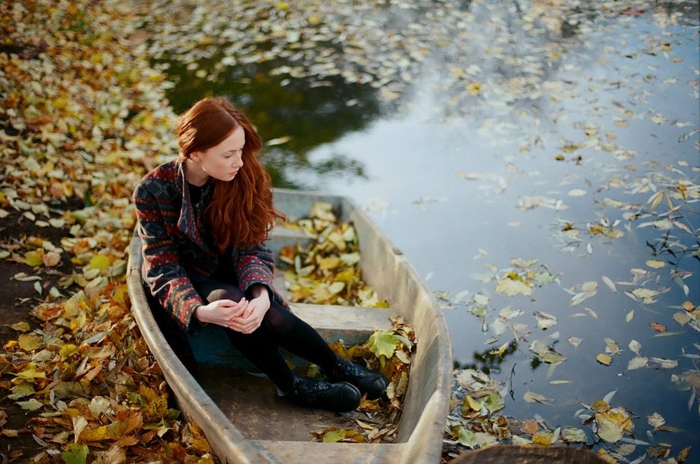 Обои для рабочего стола Девушка сидит в лодке причалившей у берега реки. Вода, берег и лодка усыпаны осенними листьями, фотограф Ксения Засецкая