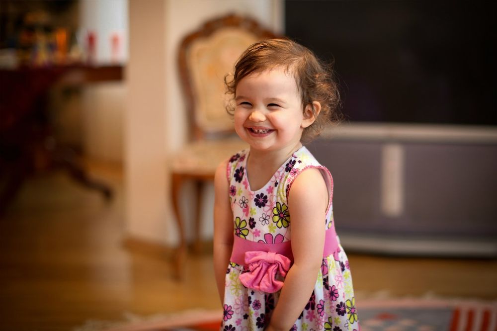 Обои для рабочего стола Маленькая смеющаяся девочка, в цветастом платье, стоит в комнате на размытом фоне, фотограф Максим Николаев