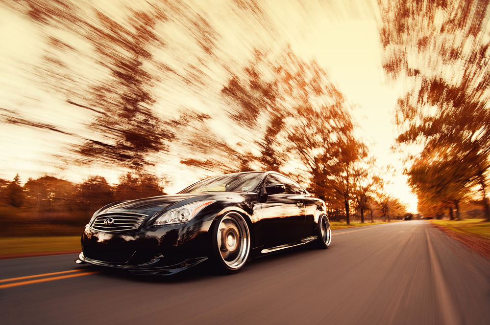 Обои для рабочего стола Автомобиль марки Infiniti G37 Coupe, мчит по дороге на фоне размытых деревьев с желтой листвой