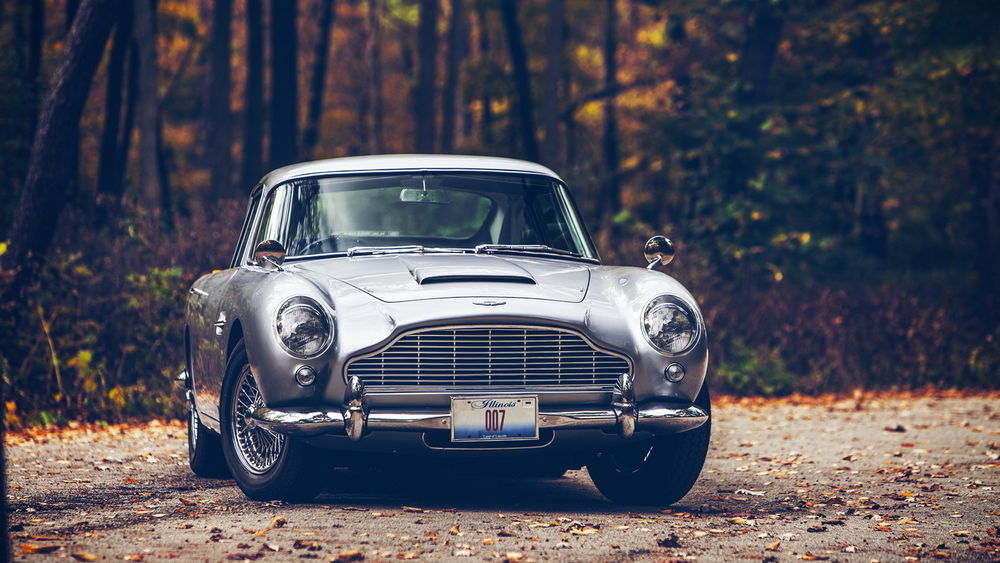 Обои для рабочего стола Серебристый автомобиль марки Aston Martin, стоит на дороге осыпанной желтыми листьями на фоне деревьев