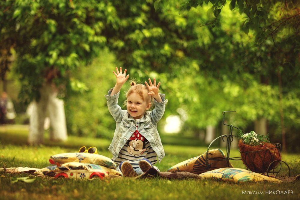 Обои для рабочего стола Рыжеволосая, улыбающаяся девочка сидит на траве подняв руки вверх, рядом лежат подушки, игрушечная машинка, солнцезащитные очки и корзинка с цветами в виде велосипеда, фотограф Максим Николаев