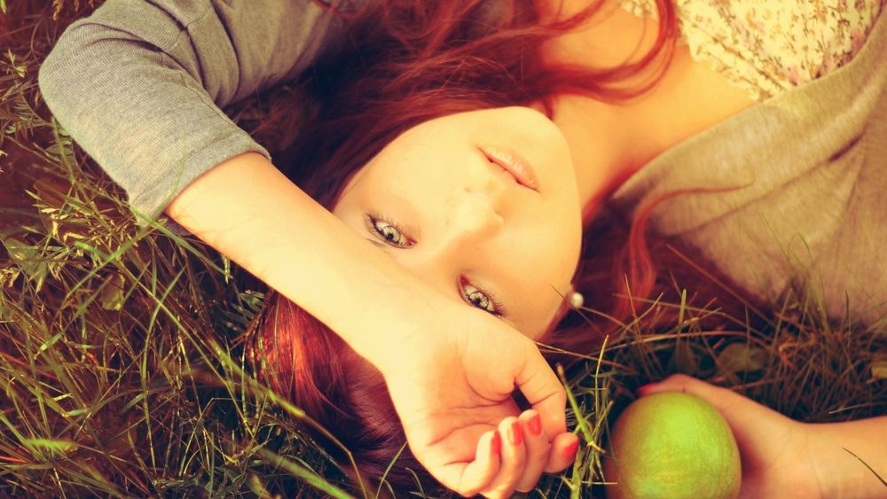Обои для рабочего стола Девушка лежит в траве, одну руку положила на лоб, в другой, держит фрукт