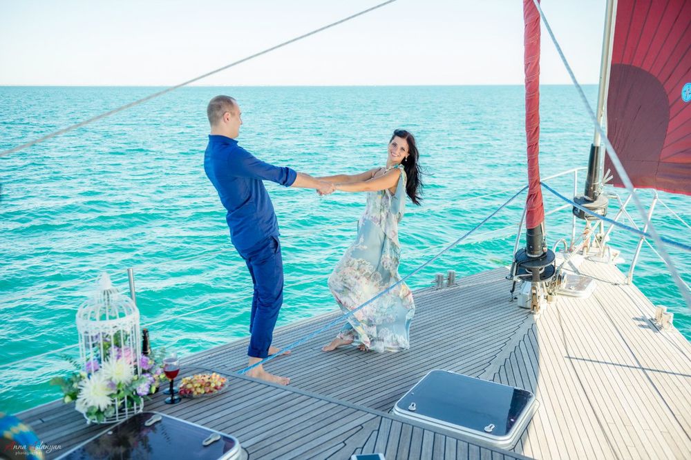 Обои для рабочего стола Девушка и мужчина, держась за руки, стоят на палубе яхты, которая вышла в море, рядом с ними цветы, бокалы с вином и еда, фотограф Анна Асланян