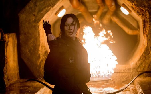 Обои на рабочий стол Дженнифер Лоуренс / Jennifer Lawrence в роли Китнисс Эвердин стоит спиной к огню с луком в руках (кадр из фильма Голодные игры: Сойка-пересмешница. Часть 2 / The Hunger Games: Mockingjay - Part 2)