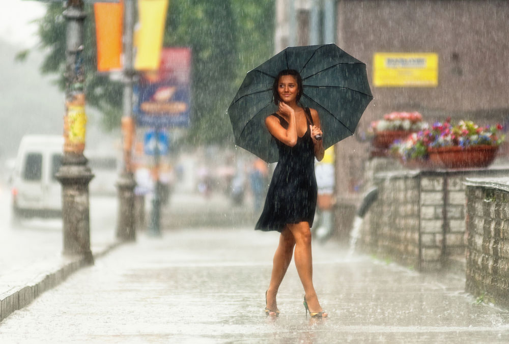 Обои для рабочего стола Счастливо улыбающаяся девушка идет под слепым летним дождиком по улице мимо корзин цветов