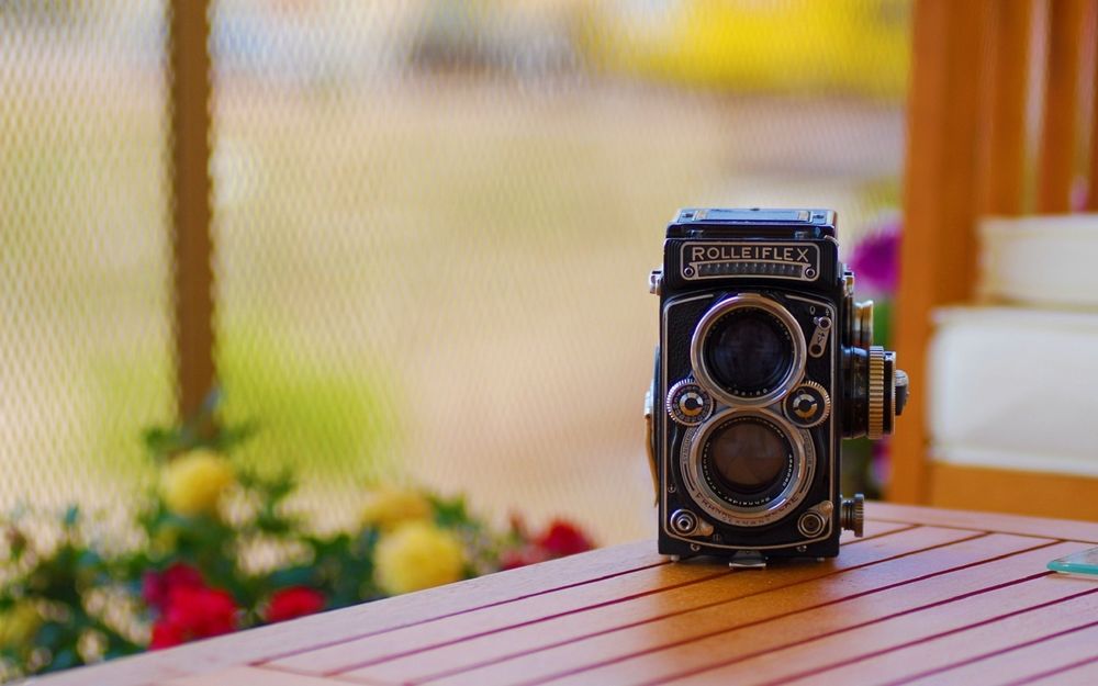 Обои для рабочего стола Двухобъективный зеркальный фотоаппарат Rolleiflex 2. 8F стоит на столе на веранде, позади, яркие цветы