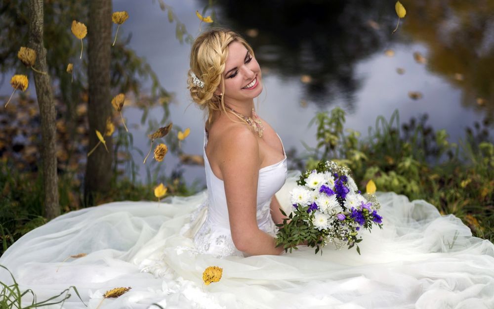 Обои для рабочего стола Девушка в свадебном платье с букетом цветов сидит на траве, у воды