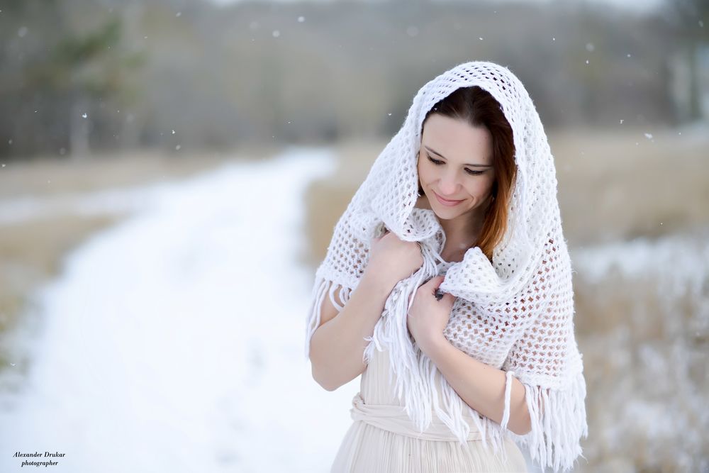 Обои для рабочего стола Улыбающаяся девушка в белом платье с белой накидкой на голове стоит среди падающего снега на дороге покрытой снегом, фотограф Александр Друкар