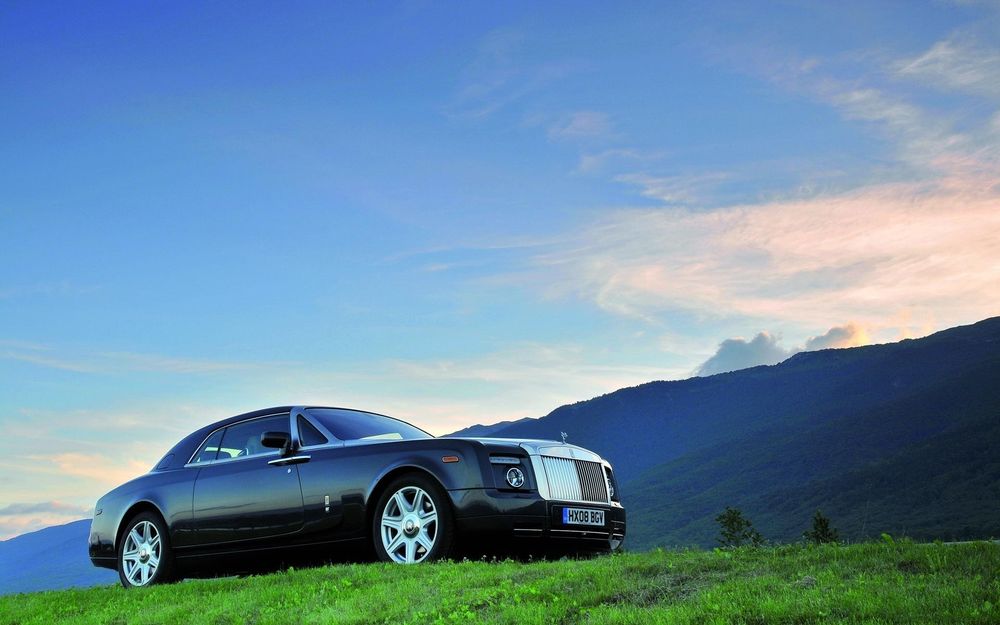 Обои для рабочего стола Автомобиль Rolls-Royce Drophead Coupe стоит на зеленой поляне на фоне гор