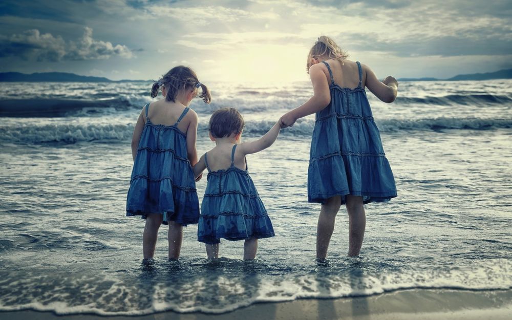 Обои для рабочего стола Три девочки разного возраста в одинаковых сарафанчиках, держась за руки стоят в воде на берегу моря и смотрят вниз на воду