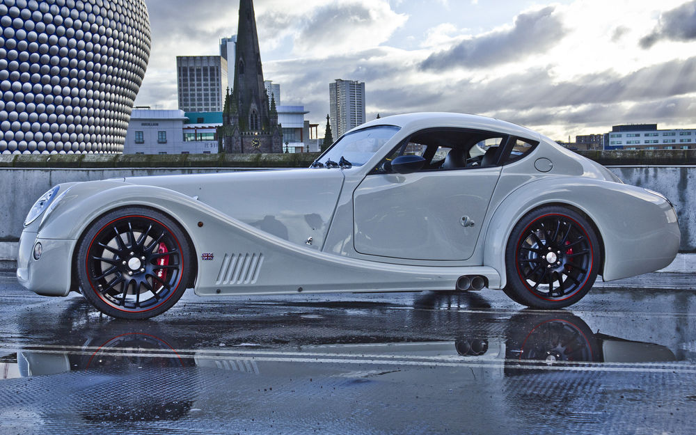 Обои для рабочего стола Автомобиль Morgan Aero Coupe стоит на мокром асфальте на фоне большого города и хмурого неба