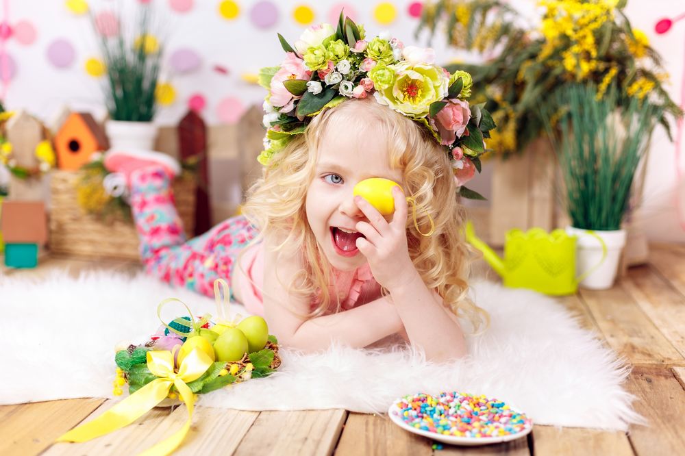 Обои для рабочего стола Девочка в веночке на голове, лежит на пушистом белом коврике и держит в руке желтое пасхальное яйцо, на фоне комнаты с цветами в горшочке