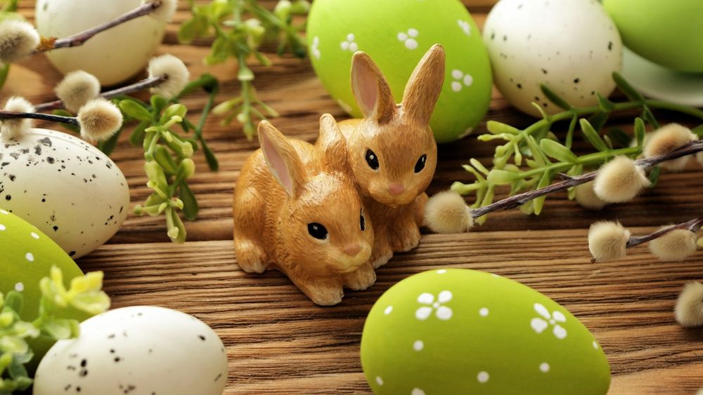 Обои для рабочего стола Статуэтки зайцев среди крашеных яиц и веточек вербы на деревянной поверхности, к празднику Светлой Пасхи