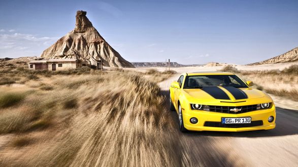 Обои на рабочий стол Желтый Шевроле / Chevrolet мчится по дороге в прерии, позади остались скала и строение