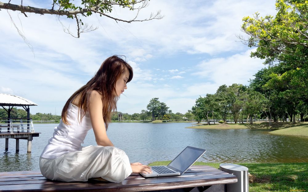 Обои для рабочего стола Девушка с ноутбуком сидит у озера, на фоне неба, водной глади и деревьев