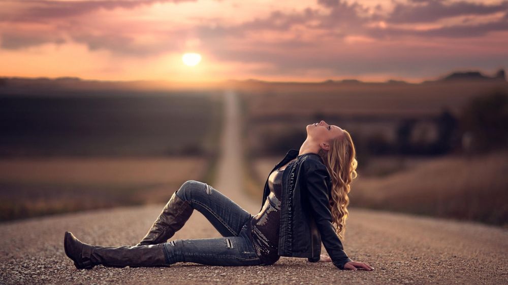 Обои для рабочего стола Девушка сидит на пустынной дороге и смотрит вверх, солнце садится за горизонт