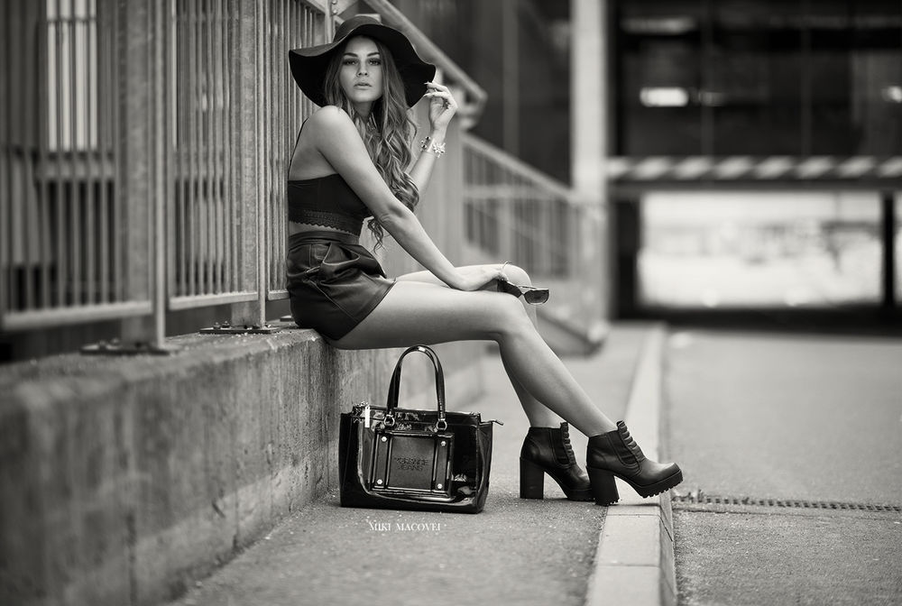 Обои для рабочего стола Девушка в кожаной юбке, топе и шляпе сидит у дороги, поставив сумку на землю, в руках у нее солнцезащитные очки, фотограф Miki Macovei