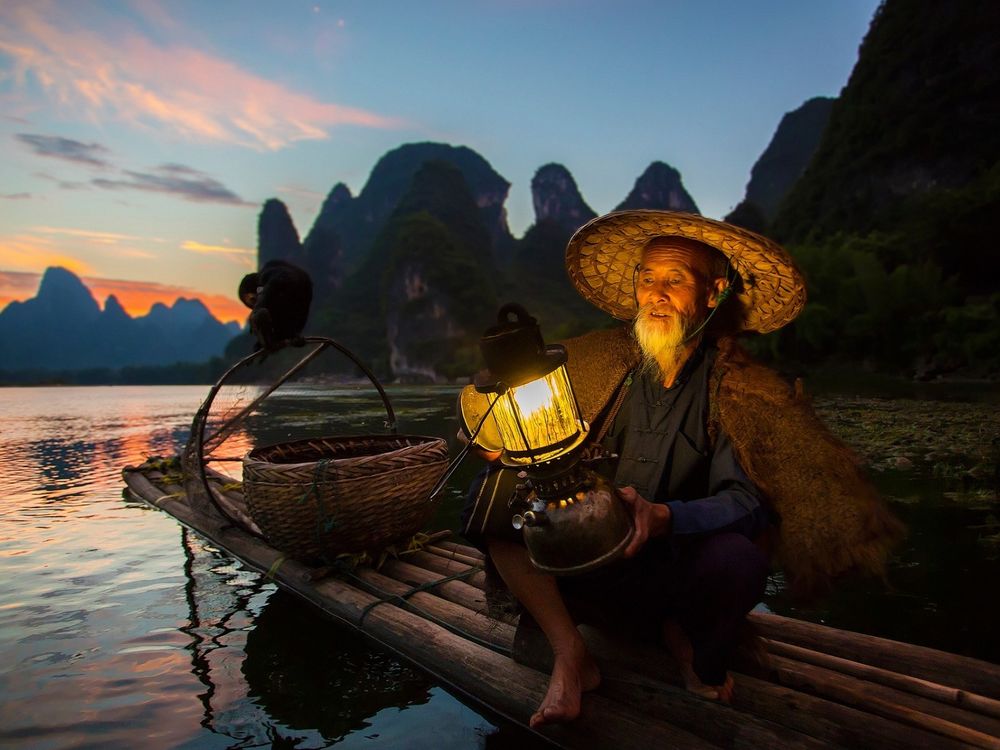 Обои для рабочего стола Старик китаец ранним утром ловит рыбу, сидя на плоту, подсвечивая себе лампой