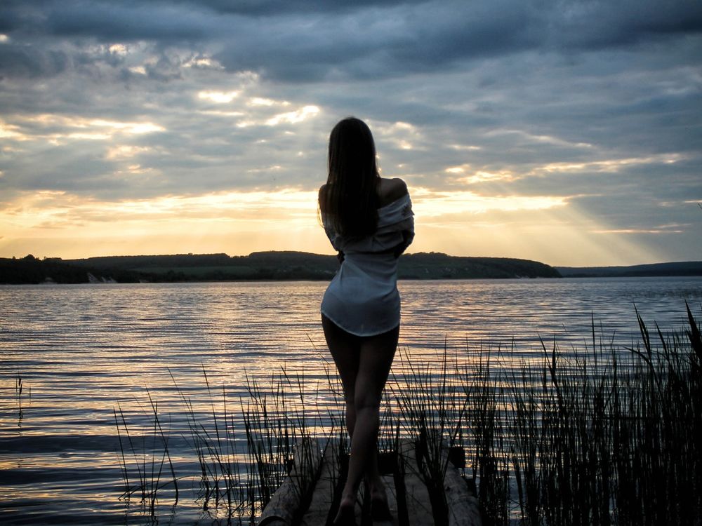 Обои для рабочего стола Длинноволосая девушка в рубахе стоит на мостках и смотрит на закат солнца над озером