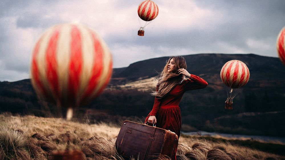 Обои для рабочего стола Девушка с чемоданом в руке стоит на поле, над которым летят большие воздушные шары