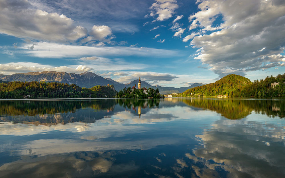 Обои для рабочего стола Озеро Блед, Словения, в воде отражается небо, вдалеке на берегу видна ратуша, фотограф Шевченко Юрий