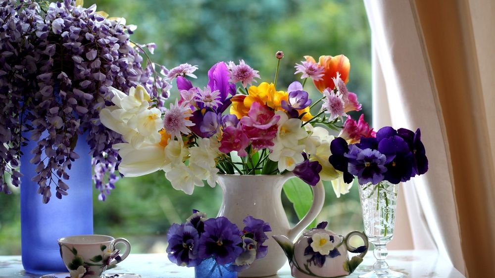 Обои для рабочего стола Разные летние цветы - глициния, анютины глазки, нарциссы, тюльпаны, хризантемы в вазах рядом с посудой в интерьере окна