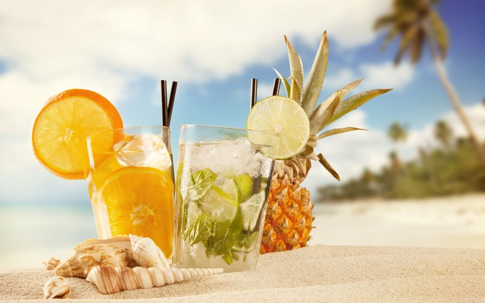 Обои для рабочего стола Два коктейля с лаймом и апельсином стоят на песке, рядом ананас и ракушки