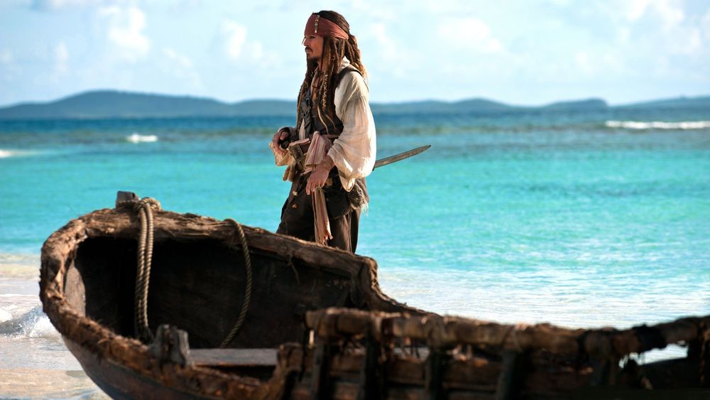 Обои для рабочего стола Капитан Джек Воробей стоит рядом с лодкой на фоне моря / кадр из фильма Пираты Карибского моря