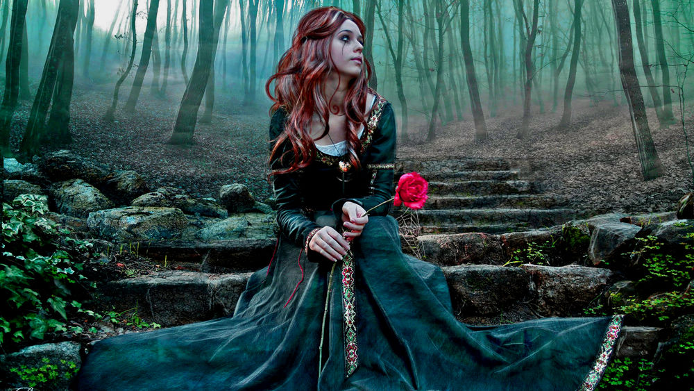 Обои для рабочего стола Рыжеволосая девушка в длинном платье, с розовой розой в руке сидит на старых каменных ступенях в лесу