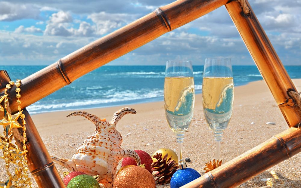 Обои для рабочего стола Два бокала с шампанским, елочные игрушки, огромная ракушка, шишки и рамка для фото из бамбука на морском берегу, в песке