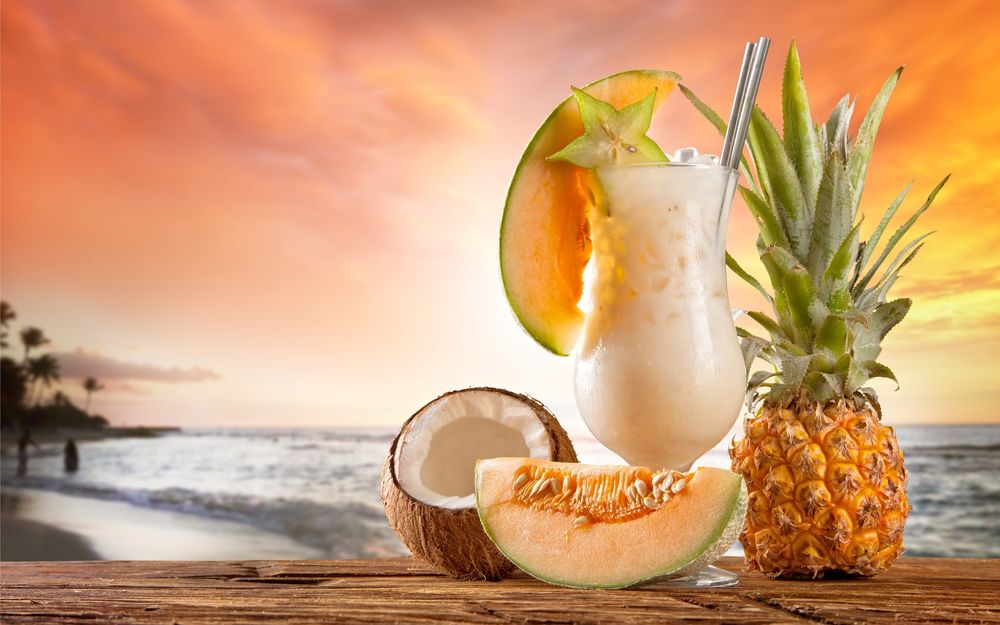 Обои для рабочего стола Тропический коктейль в высоком бокале, дыня, кокос, фрукты на деревянной поверхности, на фоне размытых неба и моря, закат солнца