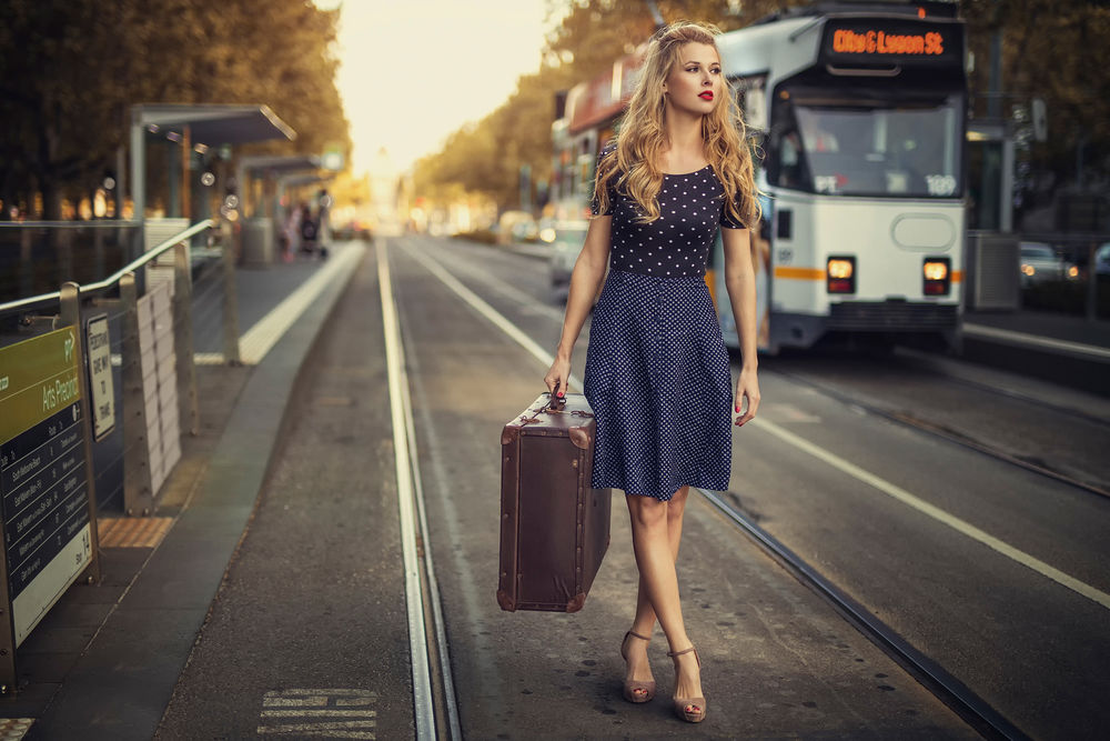 Обои для рабочего стола Девушка с винтажным чемоданом в руке стоит на городской улице в ожидании трамвая